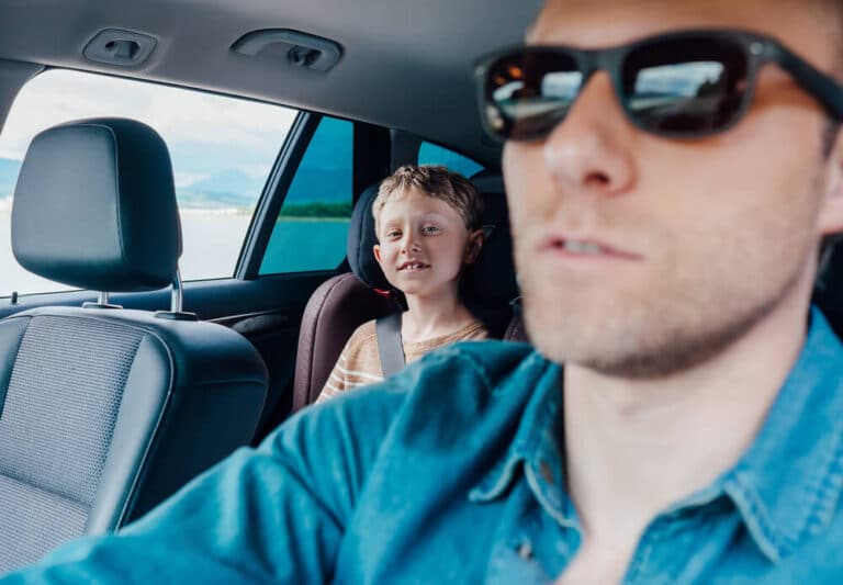 Father driving a car with son in the back seat, both enjoying a scenic view.