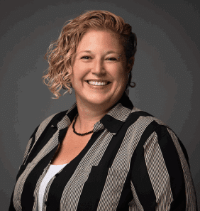 Woman with curly hair wearing a black and white striped shirt, smiling at the camera against a gray background.